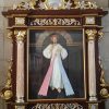 A three-dimensional mural of the Sacred Heart of Jesus shows Jesus in white clothing with white and pink rays emanating from his chest. The mural is enclosed in a brown wooden frame with glass and decorated with curly goldtone and white decorations that look like vines.