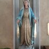 A statue of Mary inside a glass cabinet in the Shrine of Mary, Queen of Peace. She is wearing a white hood, brown robe, and blue overcoat. There's what seems to be golden rays emanating downward from both of her hands. She is also wearing a crown, and there's a halo at the back of her head. Twelve stars are attached to the halo. Her arms are down, and her palms are visible.