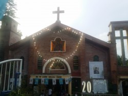 Ina ng Laging Saklolo Parish stands on a lower elevation compared to the main road, from where the picture is taken. Its facade is made of maroon bricks. It has a cross on top center, a bell tower on the left, and a tower framing a larger cross on the right. It also has rosary made of light strings between the cross and the entrance.
