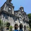 The front of San Sebastian Cathedral as seen from the corner of its yard. It has gray facade and has two towers on the sides. The curvature of the towers is due to vertical panorama shot; they are actually straight. It has several semi-oblong windows and also a semi-oblong main entrance at the center and ground level. Above the main entrance is a circular cavity with what looks like a white statue of Jesus inside. The central structure between the bell towers has a triangular roof with a cross on top. The bell towers also have crosses on top and circular windows below the bell structures.