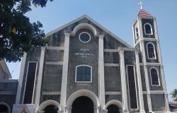 The main structure of St. James the Apostle Parish has a house-like pentagon shape and facade of that is mostly gray concrete. It has white highlights along the pillar, around the glass windows, and the semi-oblong entrances. Its entrances are one large main entrance on the ground center flanked by two slightly smaller entrances of similar shapes. It also has two vertical rectangular glass windows on the sides, one circular window on the top center, and a square window with curved top above the largest entrance. On its right (as seen by a person facing the church) is a bell tower with similar colors, three semi-oblong windows in front, one on top of another, and a red pyramidal roof with a white cross on top.