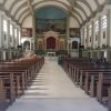 This is the nave of St. James the Apostles Parish. There are two rows of brown pews that lead to the sanctuary. It also has several rows of windows near the ceiling and rectangular wooden doors on the ground. Between the doors and the windows are murals depicting the stations of the cross.