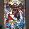 Glass Pane of St. Francis of Assisi, showing him spreading his hands upward and with his feet on the clouds. Above him are four little angels (winged heads) while aligned with his torso and feet are two more full-bodied angels: one is standing, raising one hand, and has a red cape, while the other is kneeling and praying. Below his feet is a walking angel with yellow wings, a staff, three crosses with one axis, and what seems to be a bell tower of a church.