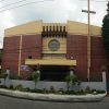 The front of San Nicolas De Tolentino Parish has a facade of red bricks in most sections of its wall. Some sections, such as those near the top and those at the center above the main entrance, are solid beige concrete. Above the main entrance, on the beige wall, is a circular glass window. A long thin beige cross is attached to the left brick wall (right of this picture). The church building is surrounded by a gray wall with an iron fence on top, making the main entrance difficult to see.